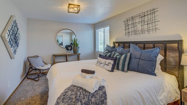 A bedroom with a large bed, a bedside table, a chair, a mirror, and a wall art piece.