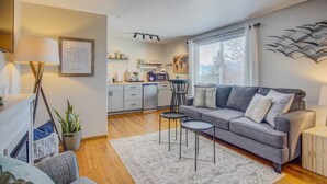 A modern living room with a grey sofa, two side tables, and a coffee table. There is a kitchen area with a microwave, cabinets, and a dining table.