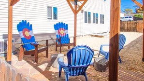 A covered patio area with wooden chairs and a table, a wooden deck, and a white house in the background.