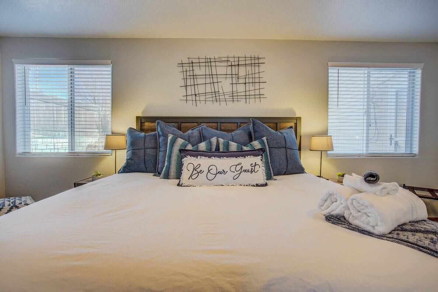 A neatly made bed with a "Be Our Guest" pillow, two bedside lamps, and a wall art piece above the bed.