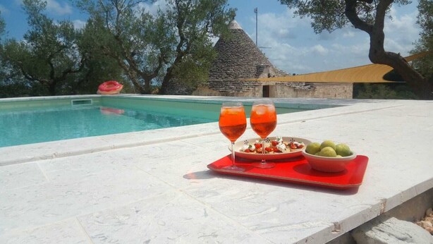 Une scène au bord de la piscine, avec une table sur laquelle se trouvent deux verres remplis d'une boisson rouge, une assiette de nourriture et un bol de fruits verts.