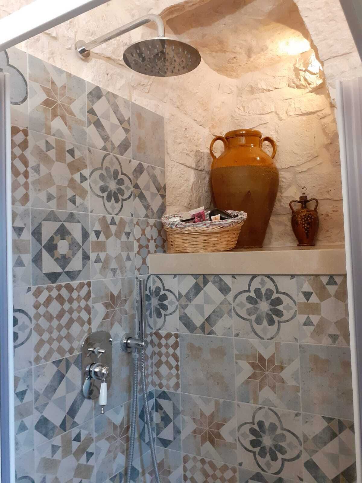 Un espace douche équipé d’une pomme de douche à effet pluie, d’un pichet en céramique et d’un panier en osier posé sur une étagère.