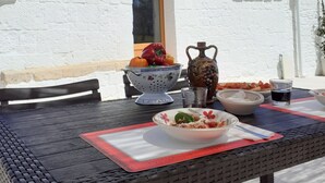 Une table avec un panier de fruits, un saladier et une carafe, le tout placé devant un bâtiment blanc doté d’un toit en pierre.