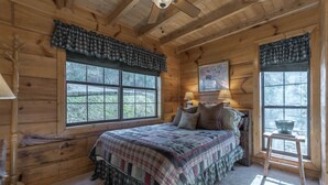 A wooden cabin bedroom with a large bed, two windows with curtains, a ceiling fan, and a lamp on a nightstand. - Private pet-friendly Aska Adventure Area getaway with gorgeous views! (Blue Ridge)