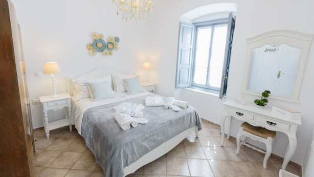 A bedroom with a bed, bedside tables, a dresser, a mirror, a chandelier, and a window with curtains. - Hydra's Pearl - Blue Pearl (Hydra)