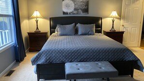 Une chambre à coucher avec un grand lit, deux tables de chevet avec des lampes, un banc, une œuvre d’art accrochée au mur et une porte.
