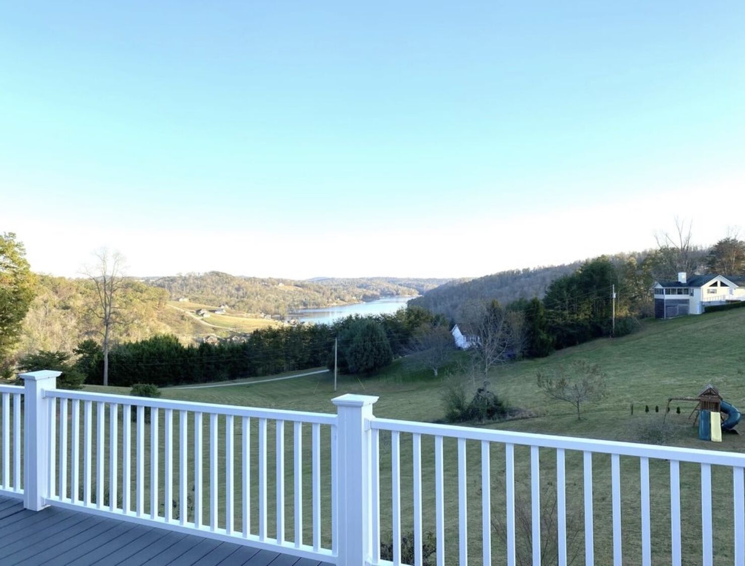 A deck with a white railing overlooking a lake, surrounded by trees and a distant view of a house.