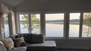 Una sala de estar moderna con un sofá seccional gris, una mesa de centro oscura y ventanas amplias que ofrecen vistas a un lago tranquilo y árboles.