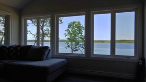 Una habitación con un amplio ventanal con vista a un lago, un sofá de color oscuro y una puerta a la derecha.