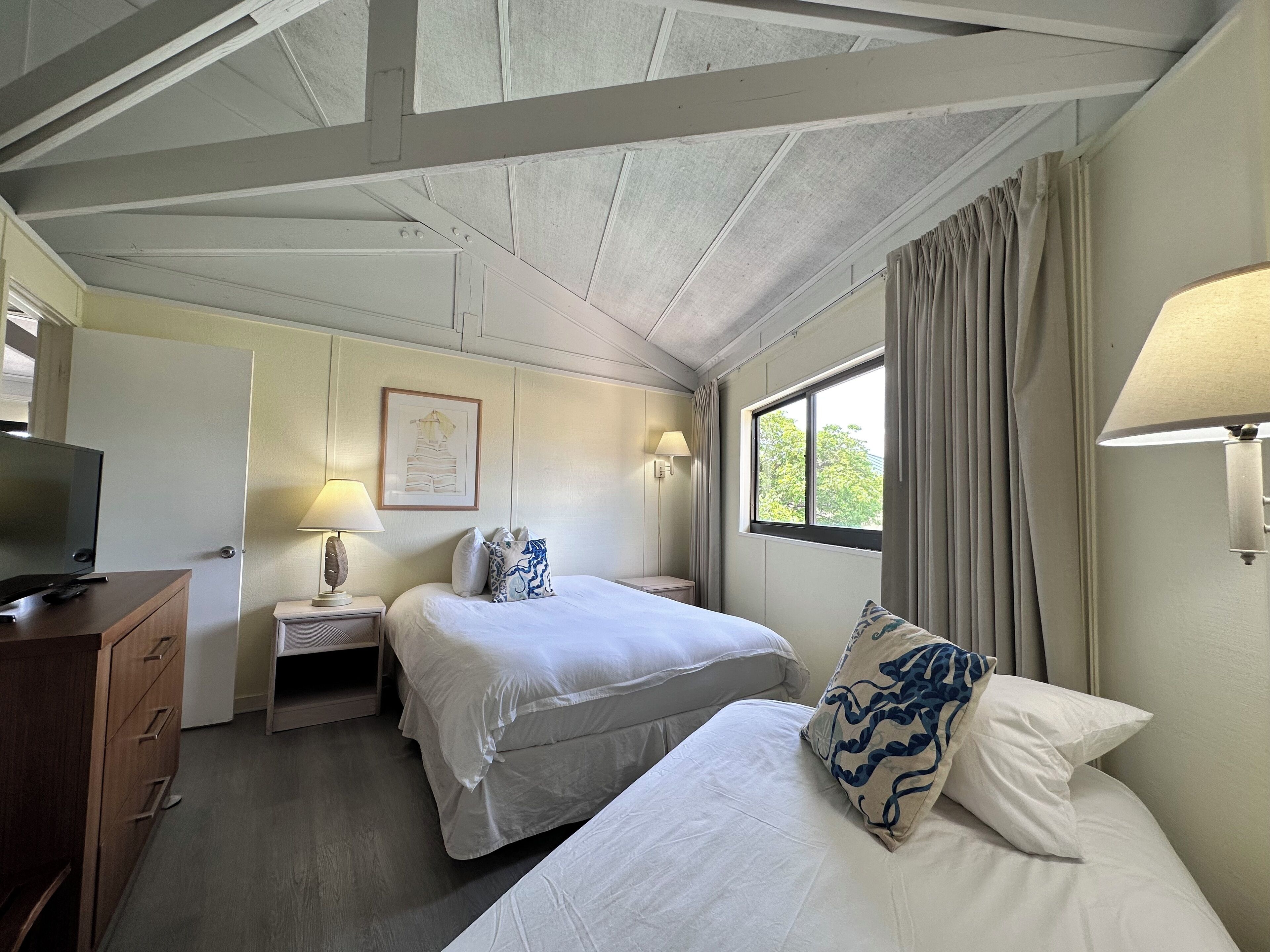 Quarto de hotel com duas camas, uma escrivaninha com televisão, uma lâmpada e uma janela com cortinas.