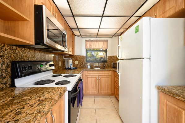 A kitchen with granite countertops, a white stove, a microwave, a refrigerator, wooden cabinets, and a window with a curtain.