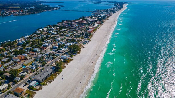 An aerial view of a coastal town with a long beachfront, clear blue waters, and a mix of residential and commercial buildings. - SURFS UP - UP STAIRS UNIT (Bradenton Beach)