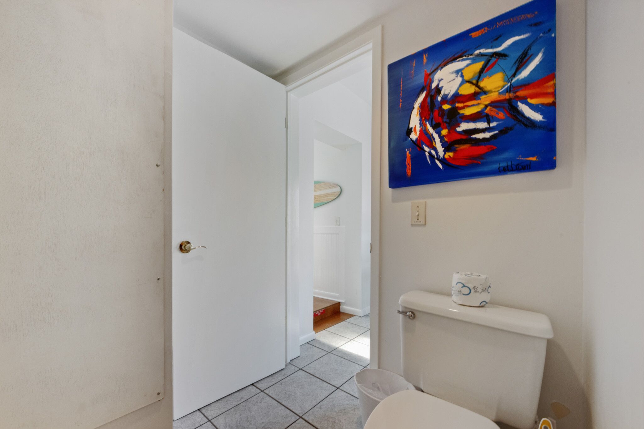 A bathroom with a white toilet, a blue abstract painting on the wall, and a white door leading to another room.