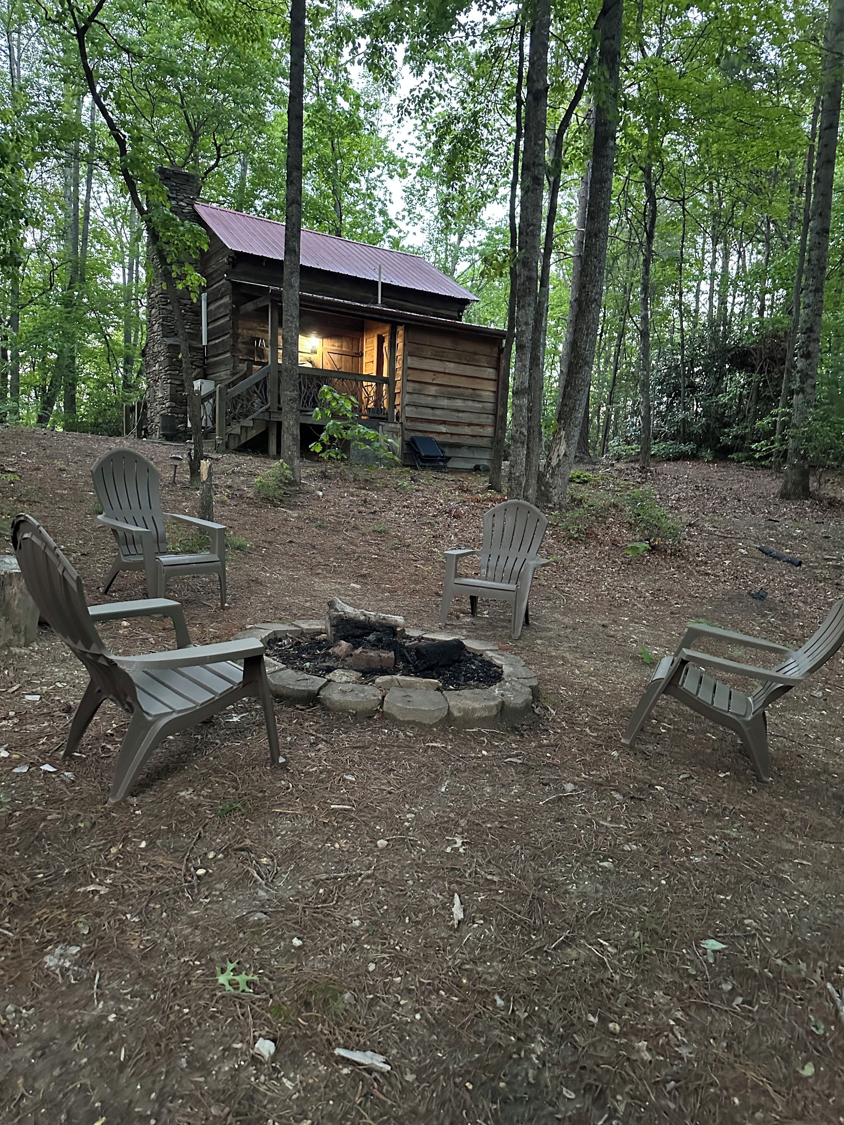 A rustic cabin with a fire pit and four Adirondack chairs in a wooded area.