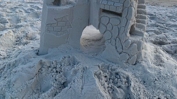 Un castillo de arena con dos torres y una pequeña entrada, ubicado en una playa con el océano al fondo.
