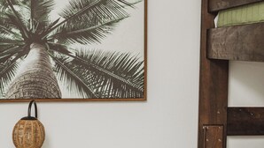 A room with a bed, a green blanket, a pillow, a wall-mounted picture of a palm tree, and a round wooden table with a lamp. - Low Rider - Bottom Unit -  (Bradenton Beach)