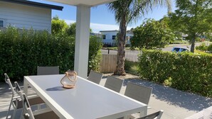 A covered patio with a white table and chairs, a glass jar with a rope handle, and a view of a residential area with a fence and trees. - Beach House (Tauranga)