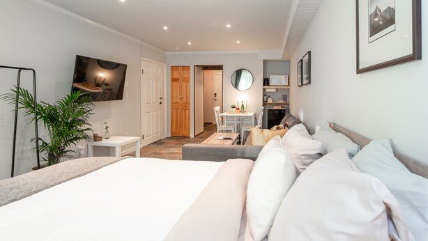 A modern hotel room with a large bed, a sitting area with a sofa and armchairs, a dining table, and a kitchen area.