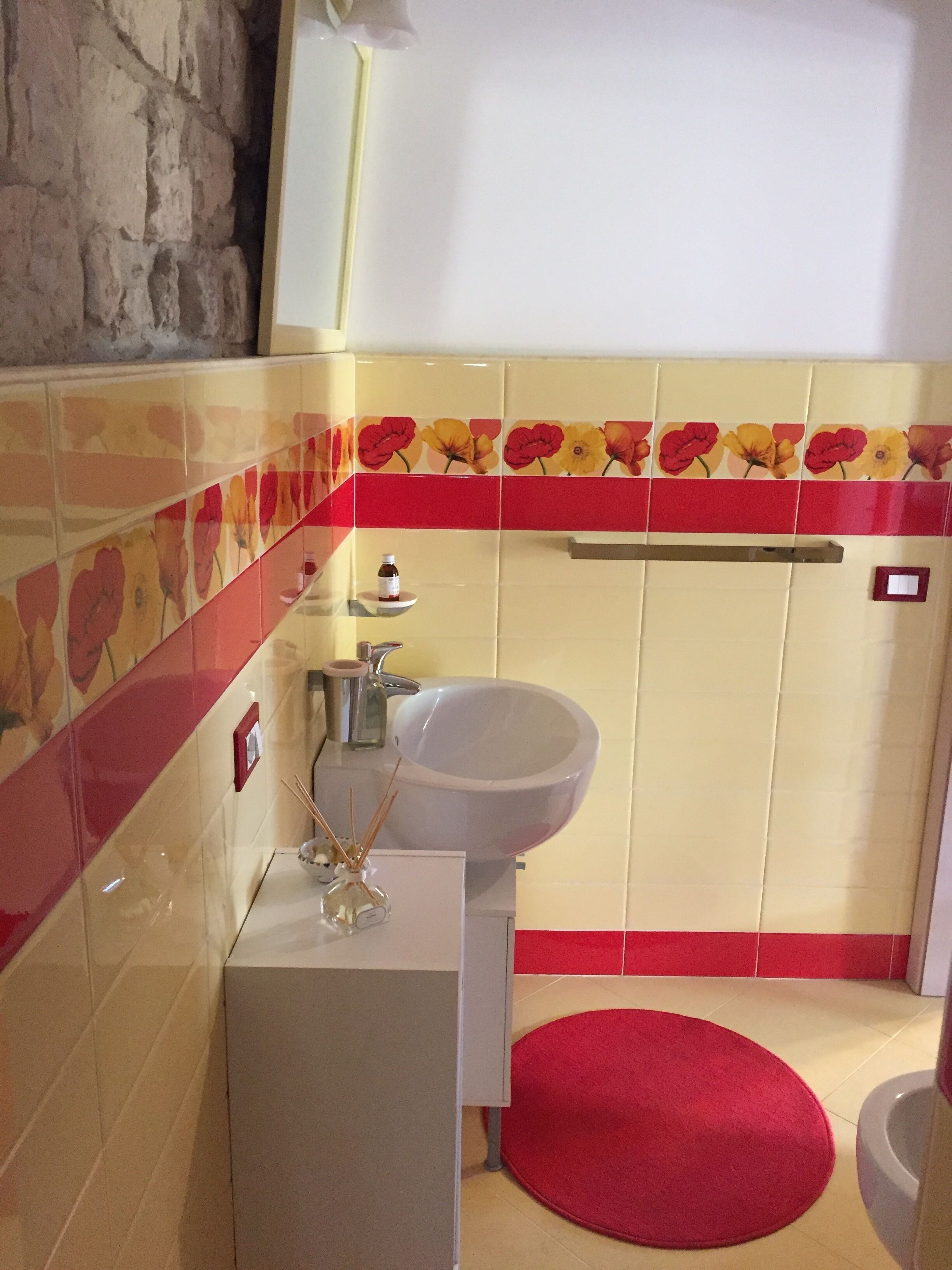 Une salle de bain avec un lavabo blanc, des carreaux de céramique aux motifs floraux rouges et jaunes, un tapis de bain rouge et un porte-serviettes.