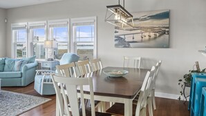 A dining area with a wooden table and white chairs, a blue sofa, and a view of a bridge through large windows. - Gorgeous Ocean and Sound Views Await at Good Better Beach! (North Topsail Beach)