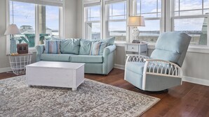 A spacious room with a light blue sofa, a white armchair, and a small white table with a lamp. Large windows offer a view of the outdoors. - Gorgeous Ocean and Sound Views Await at Good Better Beach! (North Topsail Beach)