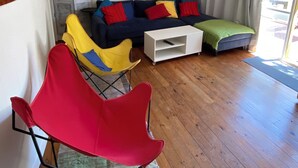 A living room with a blue and yellow painting, a dark blue sofa with red and yellow cushions, a white coffee table, and a red chair. - Tyrone Foreshore Treetop Cottage (Rye)