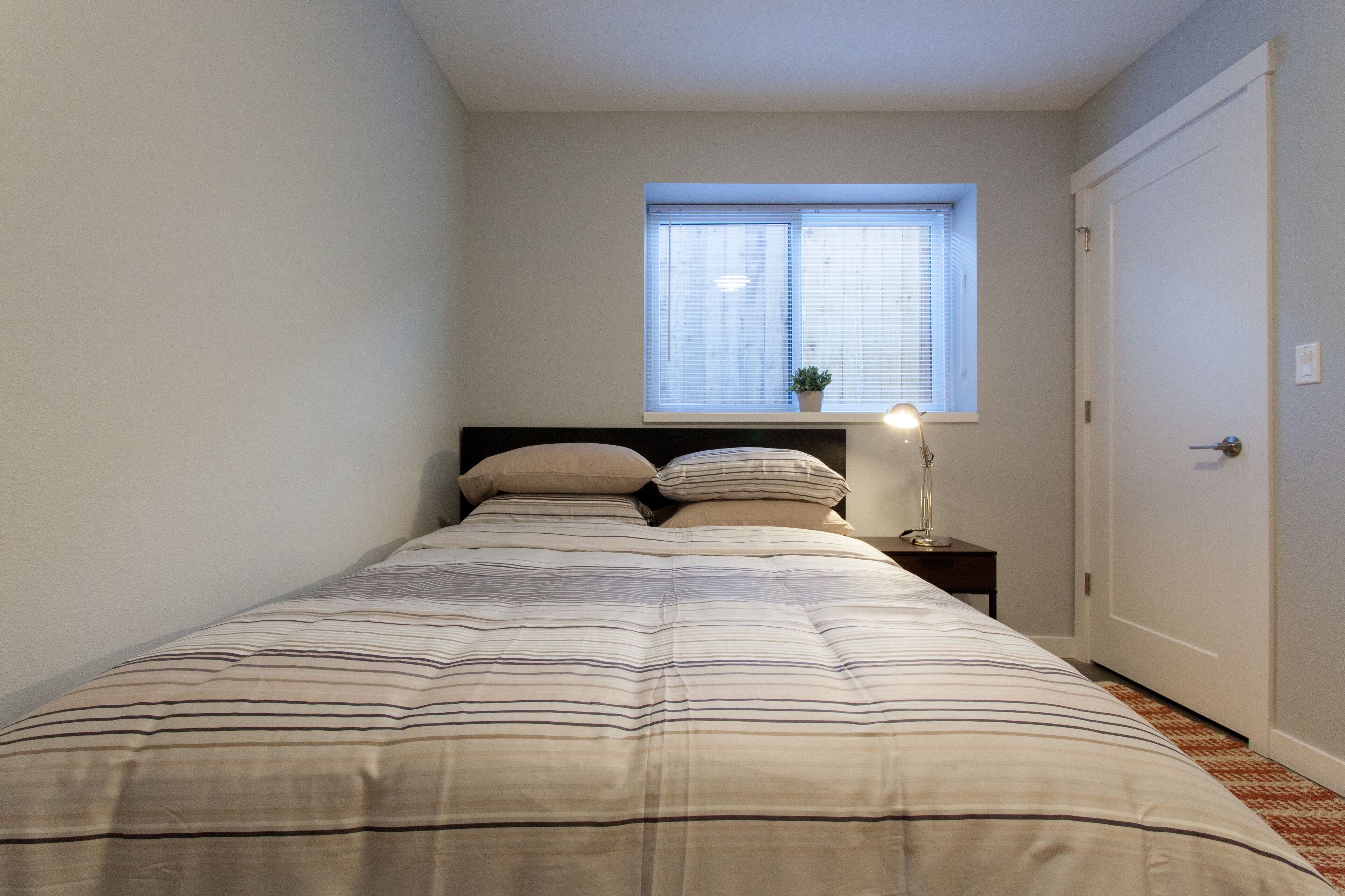 A bedroom with a bed, a nightstand, a lamp, a window, and a door.