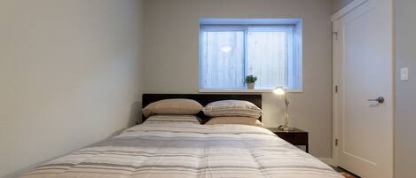 A bedroom with a bed, a nightstand, a lamp, a window, and a door.