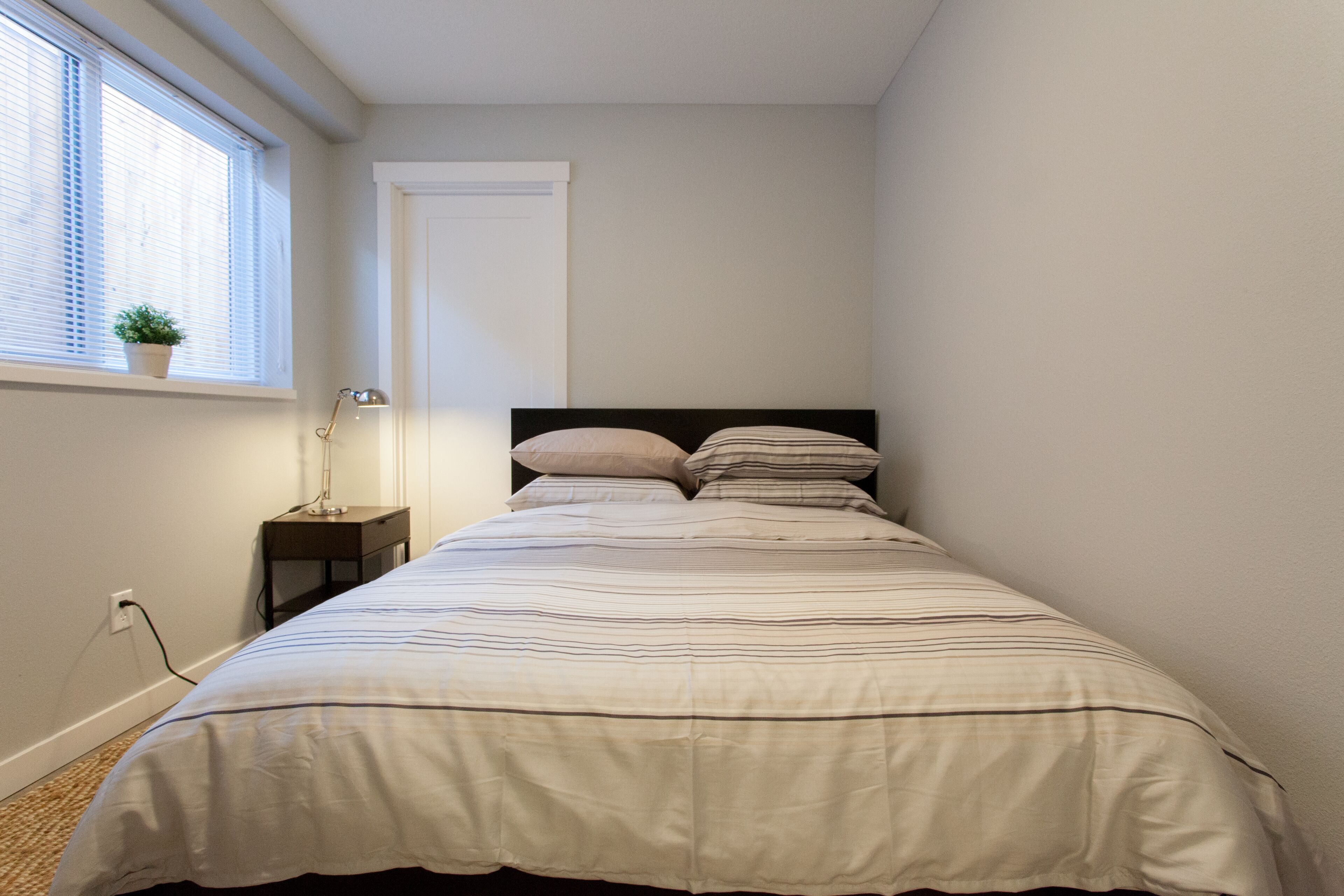 Een slaapkamer met een bed, nachtkastje, lamp, raam met zonwering en een deur.