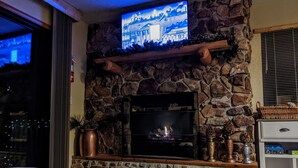 A stone fireplace with a television mounted above it, a wooden shelf with decorative items, and a bed with a cow print blanket.