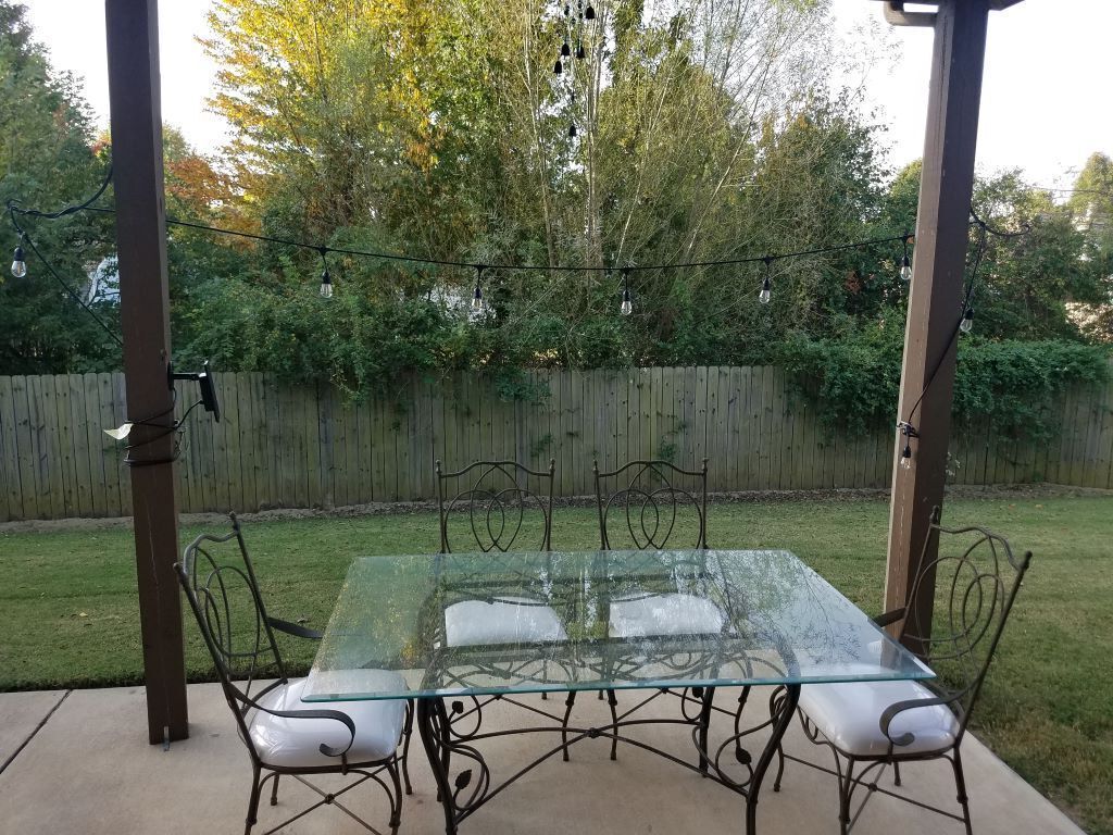 An outdoor patio with a glass-top table and metal chairs, surrounded by a wooden fence and trees.
