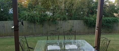 An outdoor patio with a glass-top table and metal chairs, surrounded by a wooden fence and trees.