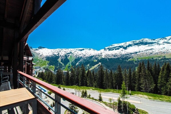 A mountain view from a balcony with a wooden railing, a paved road, and snow-capped mountains in the background.