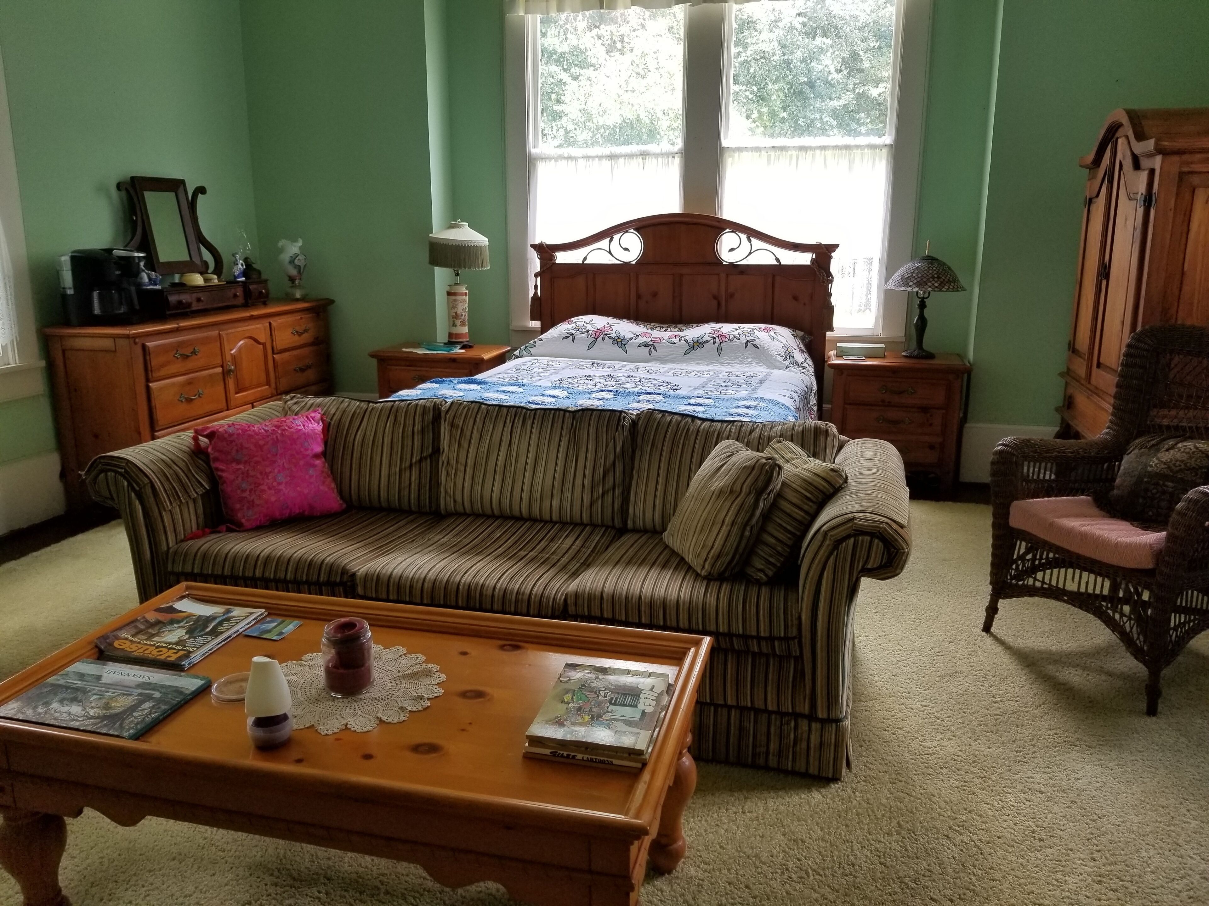 Una habitación acogedora con un sofá a rayas, una mesa de centro de madera con libros, una silla de mimbre y una cama con colcha estampada.