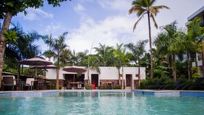 A swimming pool with lounge chairs and umbrellas, surrounded by palm trees and a building with a white facade.