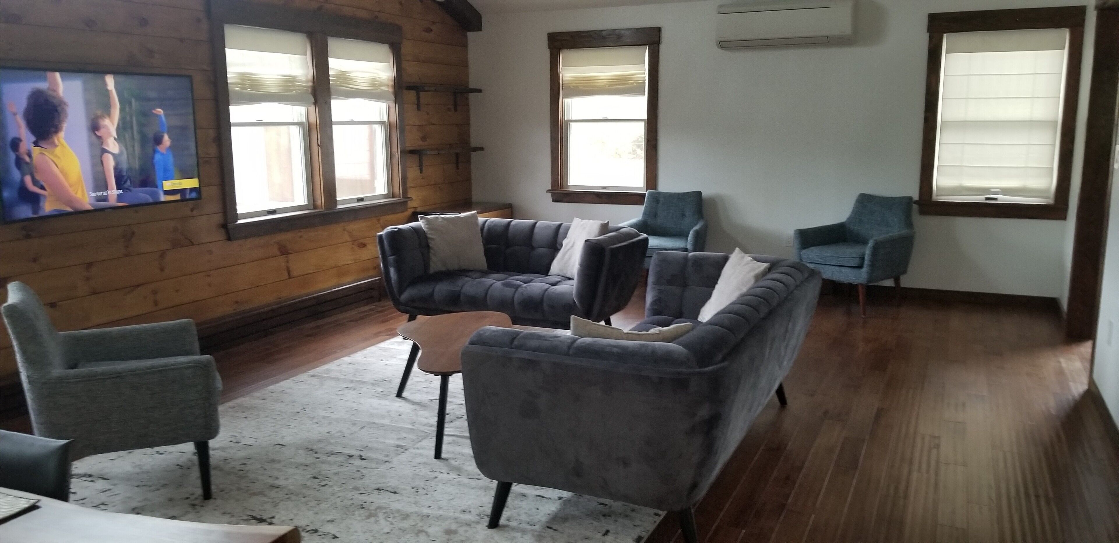 Un salon avec un canapé gris, des fauteuils, une table basse et un téléviseur fixé au mur. Il y a des fenêtres avec des stores, des murs en bois et un sol recouvert d'une moquette.