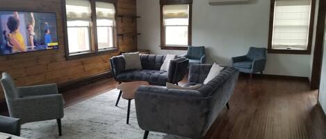 Un salon avec un canapé gris, des fauteuils, une table basse et un téléviseur fixé au mur. Il y a des fenêtres avec des stores, des murs en bois et un sol recouvert d'une moquette.