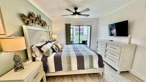 Un dormitorio con cama, mesitas de noche, cómoda, ventilador de techo, televisor y vista al exterior.