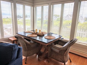 A dining area with a table set for four, surrounded by wicker chairs, with a view through large windows.