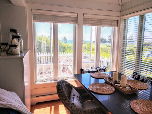 A dining area with a table set for two, a wicker chair, and a view of a garden through large windows.