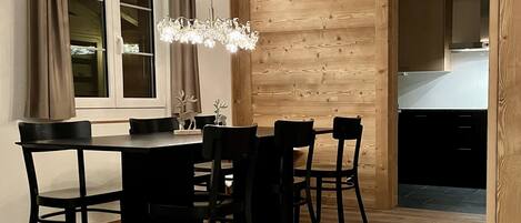 A dining area with a wooden wall, a chandelier, and a view into the kitchen.