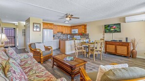 A living room with a sofa, chairs, a coffee table, a kitchen with wooden cabinets, a dining area, and a ceiling fan. - Beachside Maui condo - pool & hot tub (Kihei)