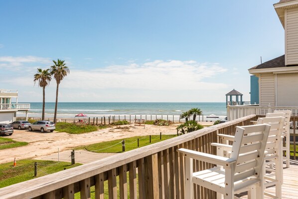 A wooden deck with white chairs overlooking a beach with palm trees and a gazebo.