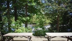 Un espace de pique-nique avec deux tables et des bancs en bois, entouré d’arbres et bordé par une rivière.