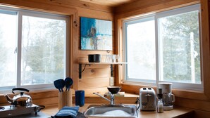 Une cuisine chaleureuse dans un chalet, avec des murs en bois, un tableau au-dessus de l’évier et une fenêtre donnant sur les arbres.