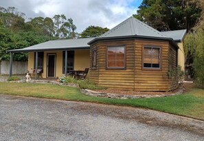 A small, single-story wooden house with a grey roof, a front porch, and a stone border around the yard. - Nowhere Else Cottage with private spa.
Unique farm stay.   (Delamere)