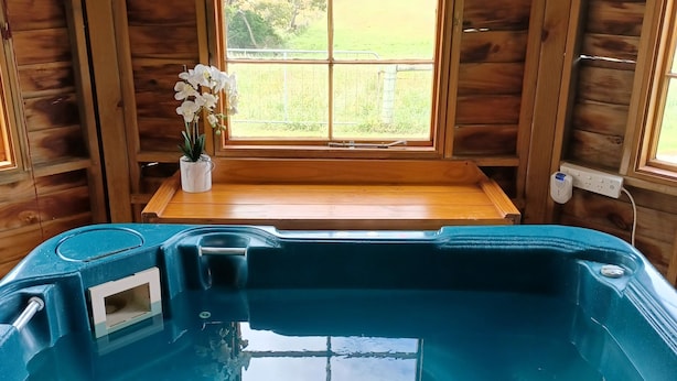 A wooden cabin with a hot tub, a window with a view of a grassy field, and a small table with a vase of flowers.