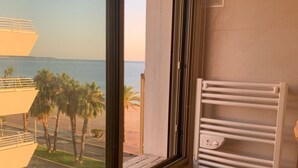 A modern bathroom with a large window overlooking a beach and palm trees.