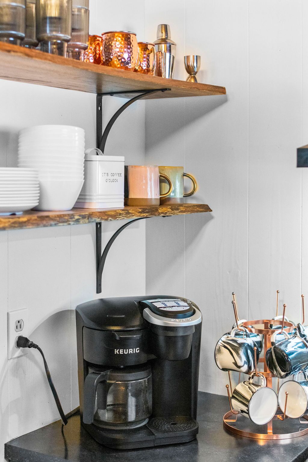 A kitchen with a Keurig coffee maker, white plates, and colorful mugs on shelves.