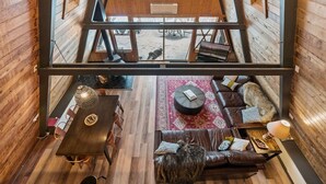 Ein gemütliches Blockhaus-Interieur mit Holzboden, einer Essecke mit Tisch und Stühlen, einer Couch mit Teppich und Blick auf eine verschneite Landschaft durch ein großes Fenster.